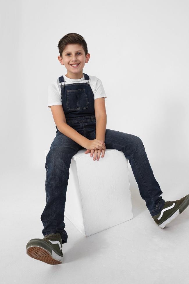 Boy sitting on box with stretched legs, wearing denim dungarees with adjustable straps.