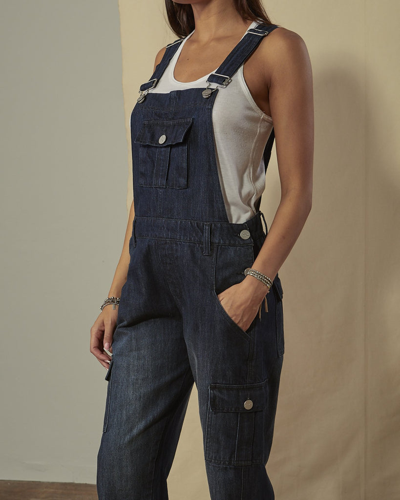 Close angled shot of women's darkwash multi-pocket bib-overalls with view of side button fastening, belt loops and adjustable straps.