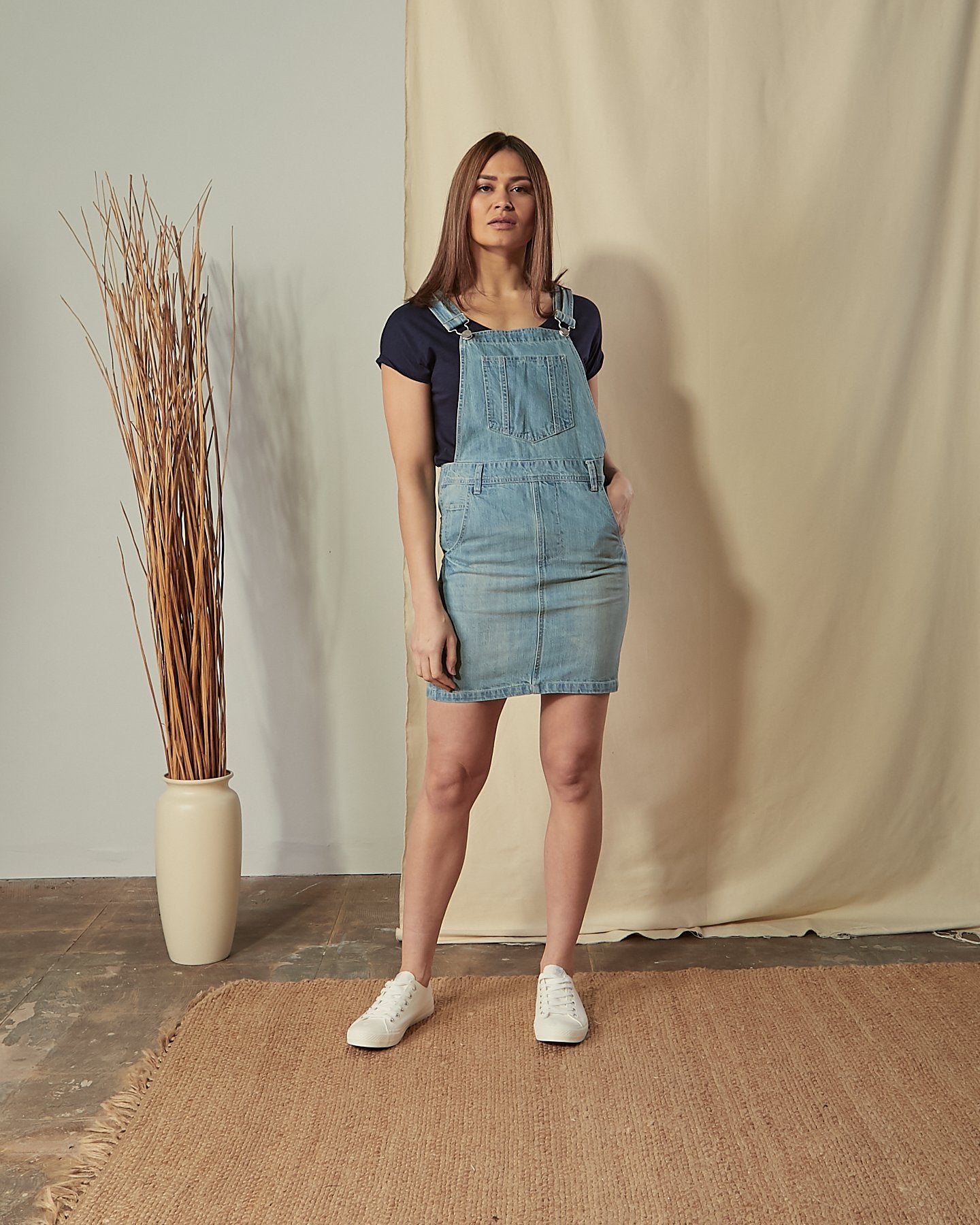Full-length front view of model with hand in front pocket of Iris relaxed fit aged aged denim dungaree dress.