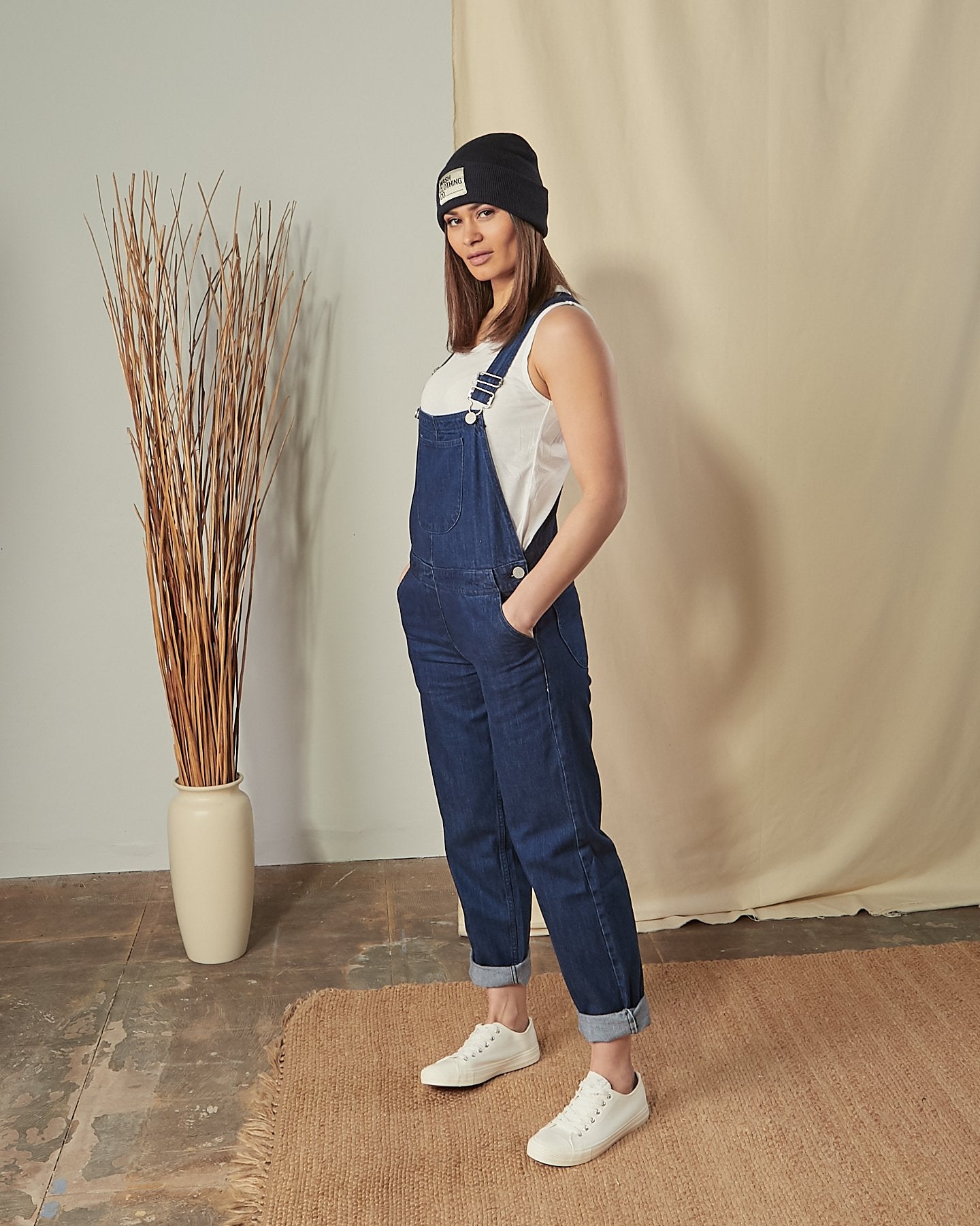 Slightly angled side view of model wearing Prue blue cotton dungarees with hands in side pockets.