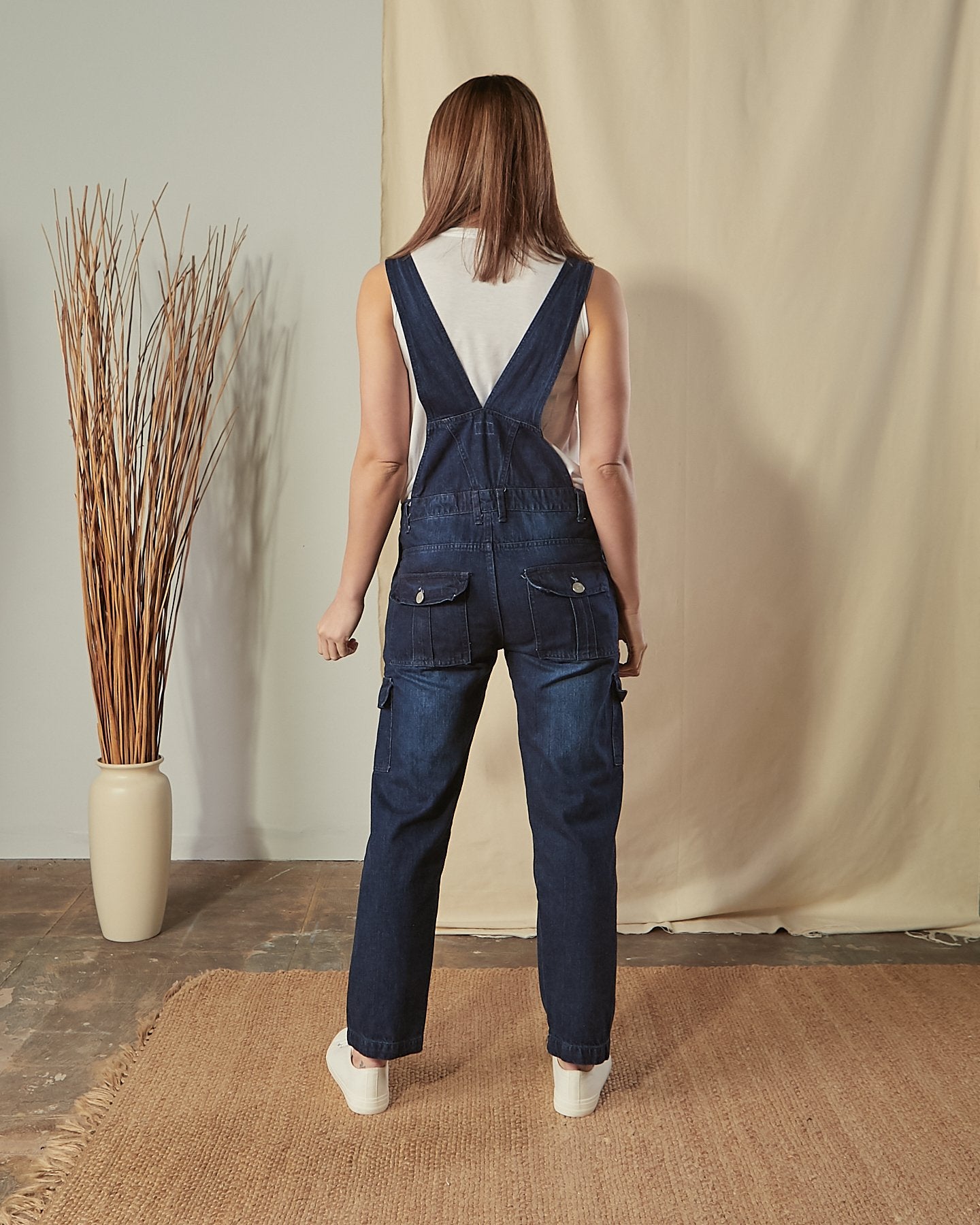 Full-length, back view of model wearing Tammy darkwash bib-overalls with ciew of belt loops, back button-down pockets. 