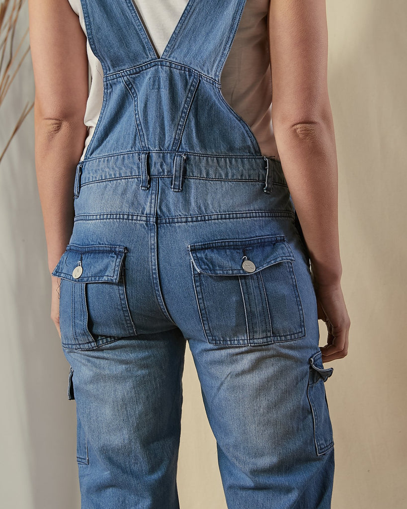 Mid-view rear of model wearing Tammy stonewash bib-overalls with view of belt loops and button-down back pockets. 