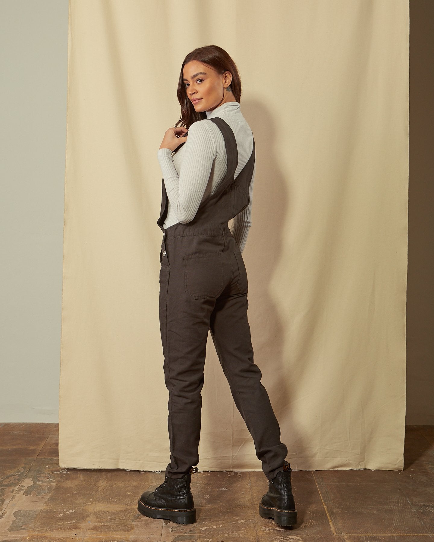 Back angled shot women's faded black organic denim bib-overalls, model looking over her shoulder with view of back pockets.