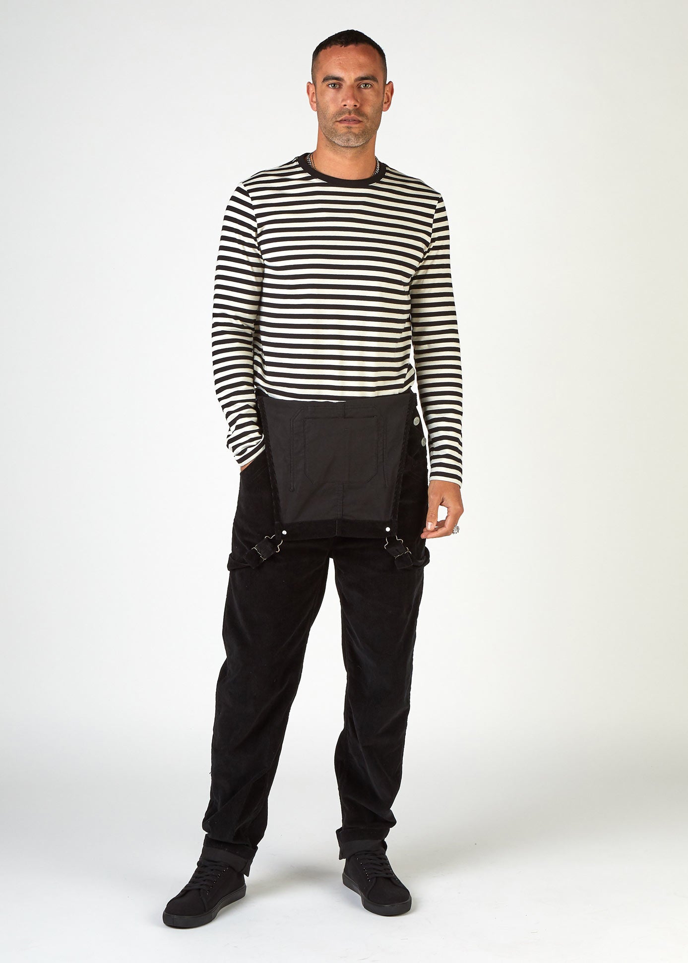 Full-length, front view of model wearing black corduroy dungarees with bib down and hand in front pocket.