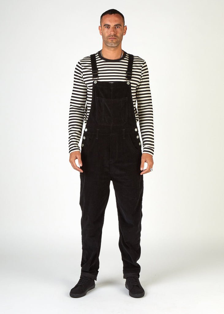 Full-length, front view of model wearing black corduroy dungarees with clear view of side button fastenings, front and bib pockets. 