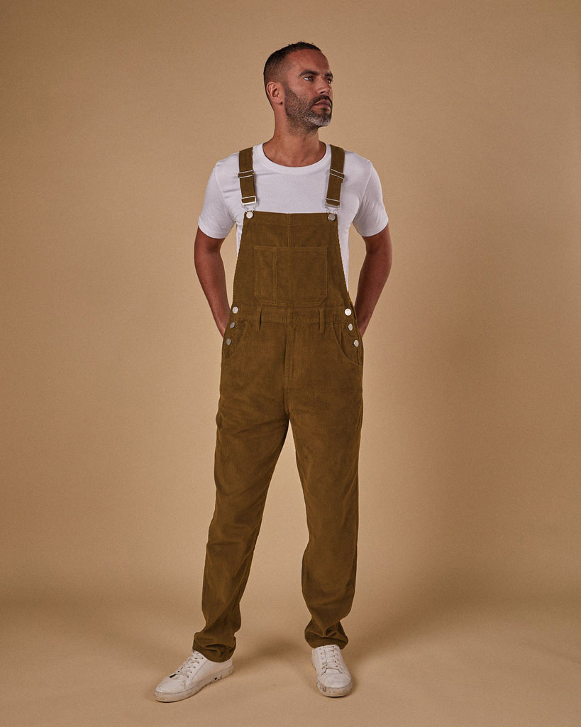 Full-length, front view of model wearing brown corduroy dungarees with clear view of side button fastenings, front and bib pockets. 