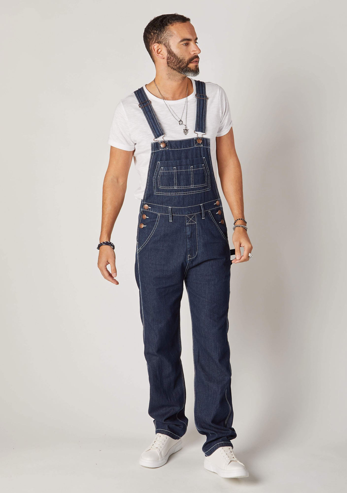 Front pose looking to left wearing loose fitting, dark blue denim bib overalls.