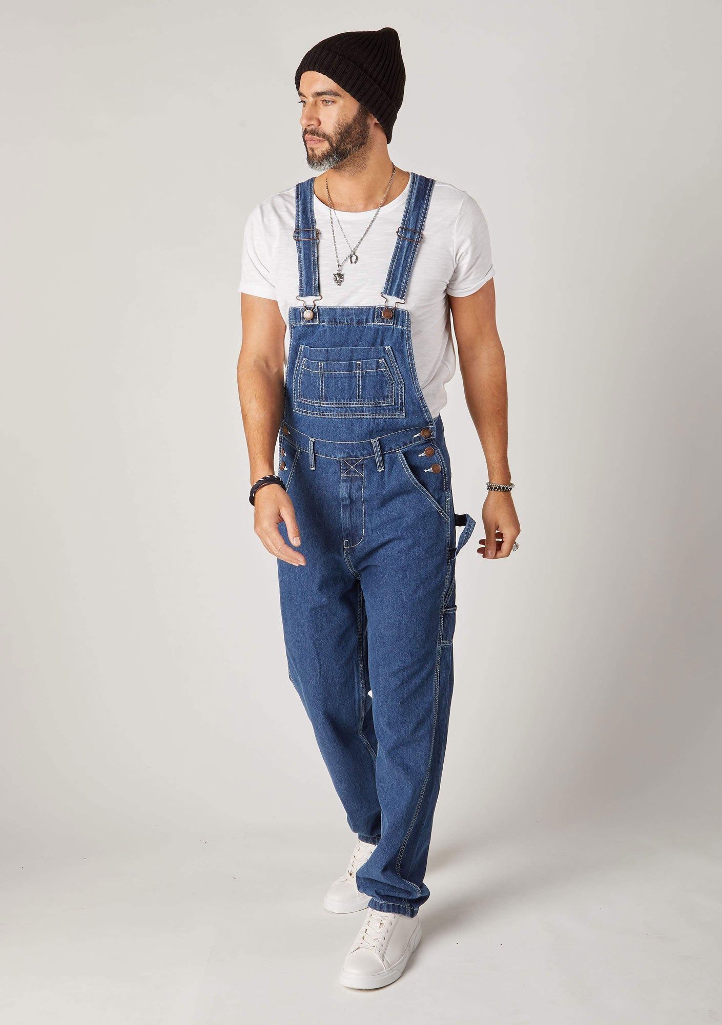 Front pose looking to left wearing loose fitting, stonewash denim bib overalls with view of bib and front pockets and adjustable straps.