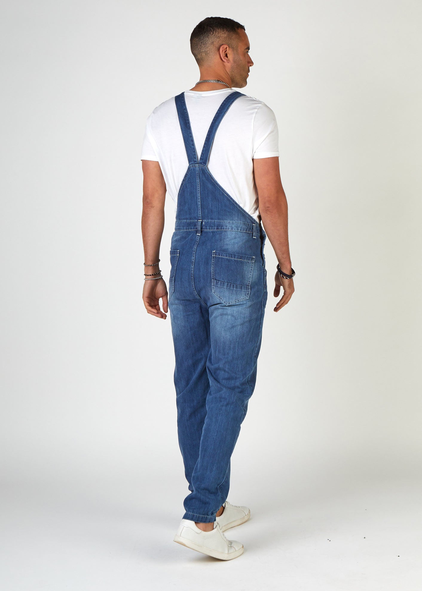 Rear full-length pose looking right, wearing Bertie-style, denim bib overalls with view of back straps and pockets.