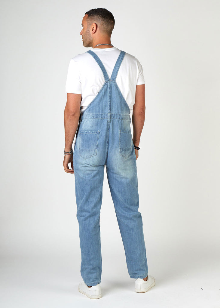 Rear full-length pose looking left, wearing Bertie-style, ripped palewash denim bib overalls with view of back straps and pockets.
