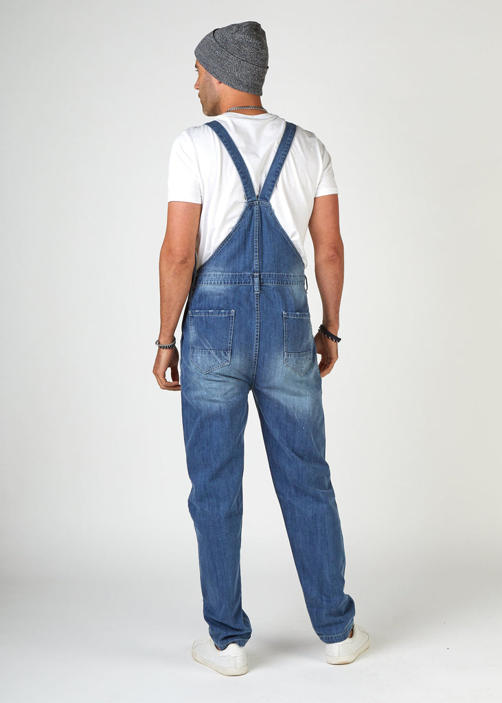 Rear full-length pose looking left, wearing Bertie-style, ripped denim bib overalls with view of back straps and pockets.