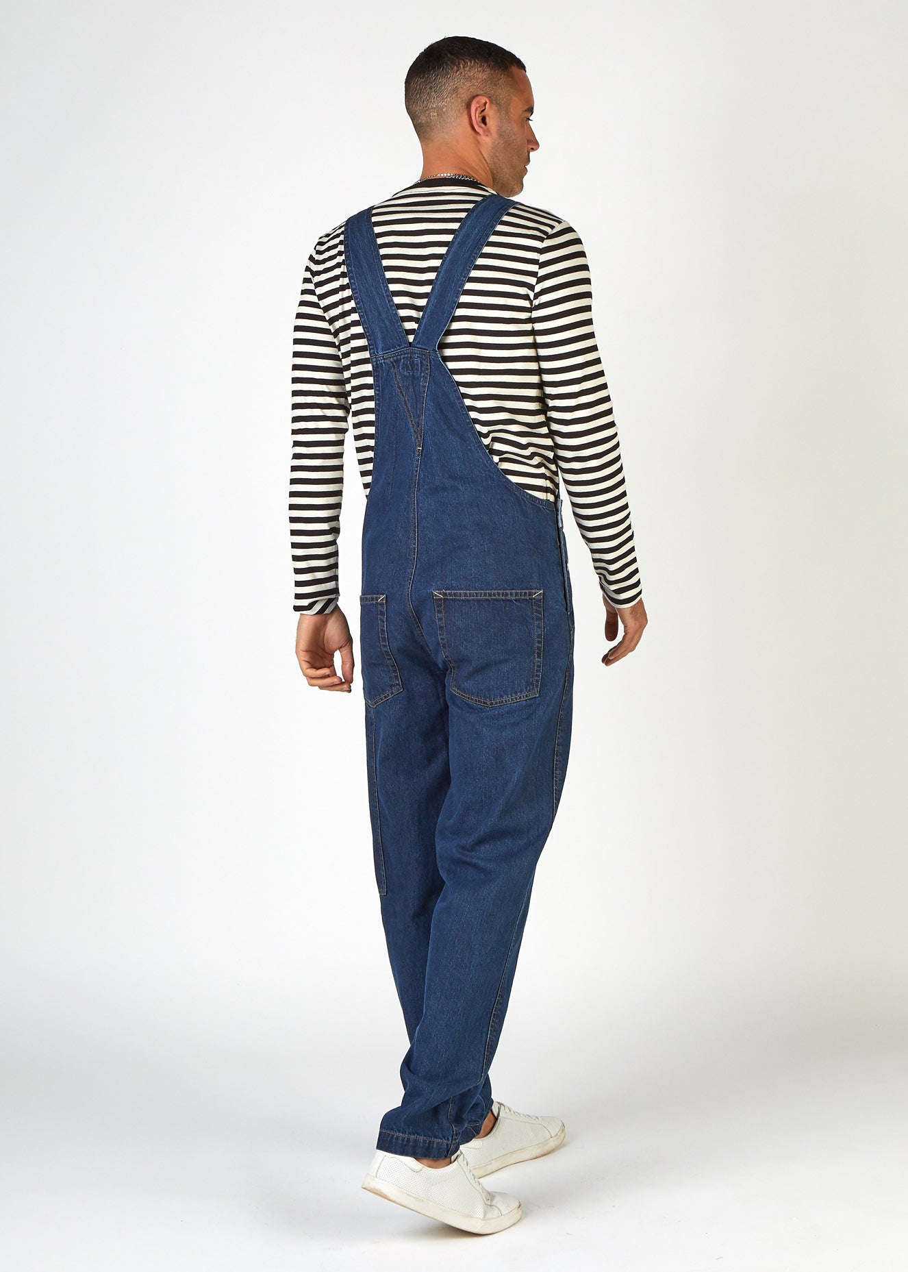 Back view twisting angled lightly to right, wearing indigo bib-overalls with clear view of back pockets and back strap.