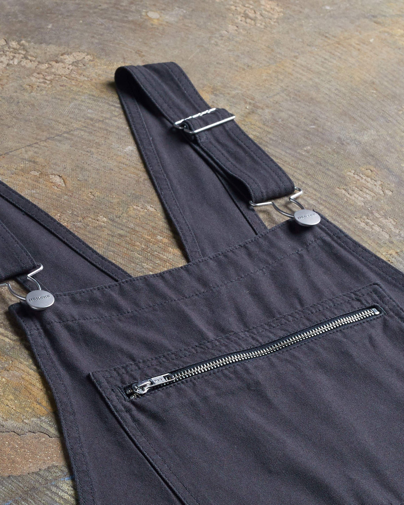 Close-up angled flat shot of organic denim bib overall dress with focus on zip-up bib pocket and adjustable straps.