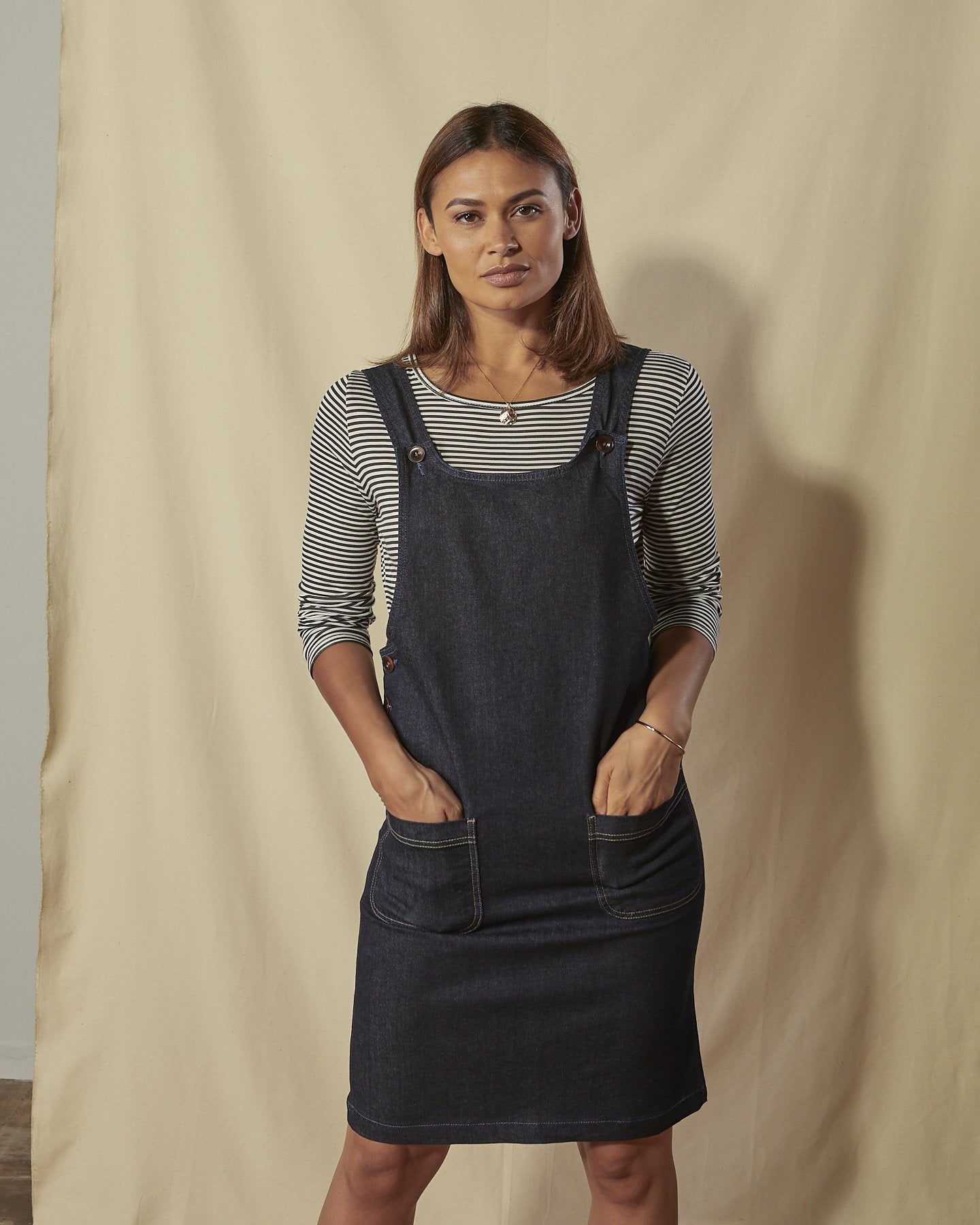 Three quarter view of Addie organic denim pinafore with view of front pockets and dungaree style front.