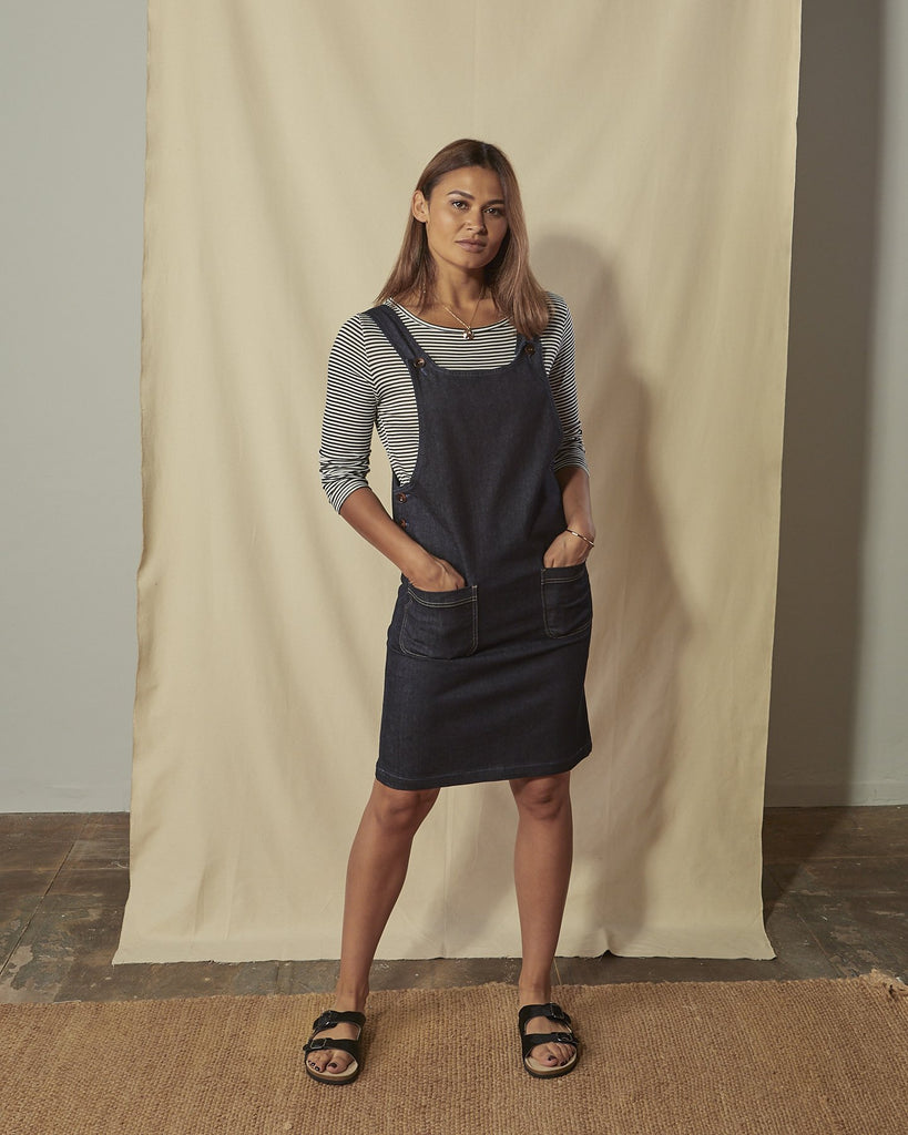 Full-length front view of model wearing sleeveless Addie organic denim pinafore with hands in front pockets.