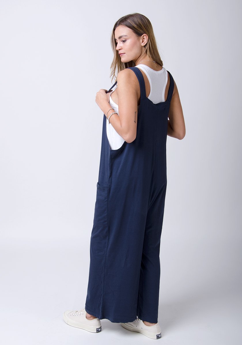 Side-rear full-length pose with Amber-style, navy, cotton jersey, relaxed-fit overalls holding adjustable straps.