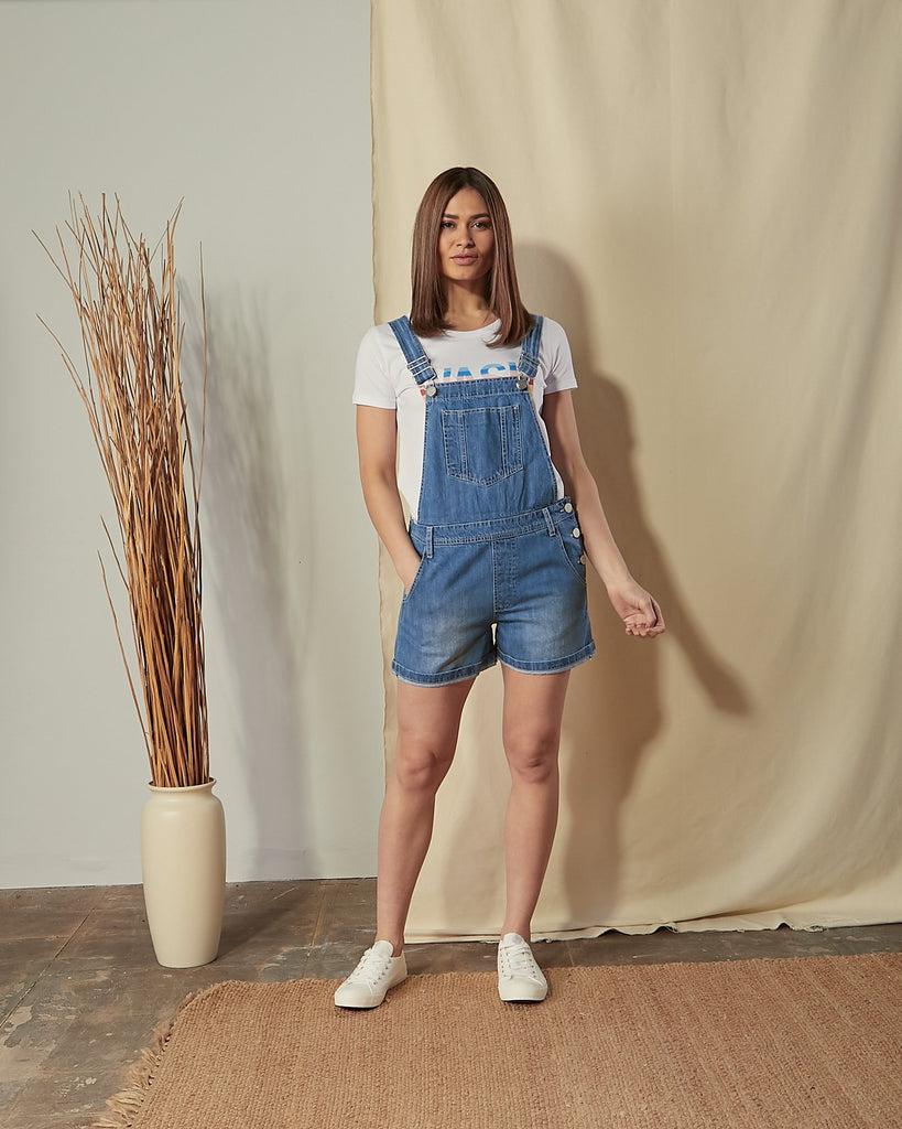 Full-length front view of model with hand in front pocket of Ava relaxed fit denim dungaree shorts.