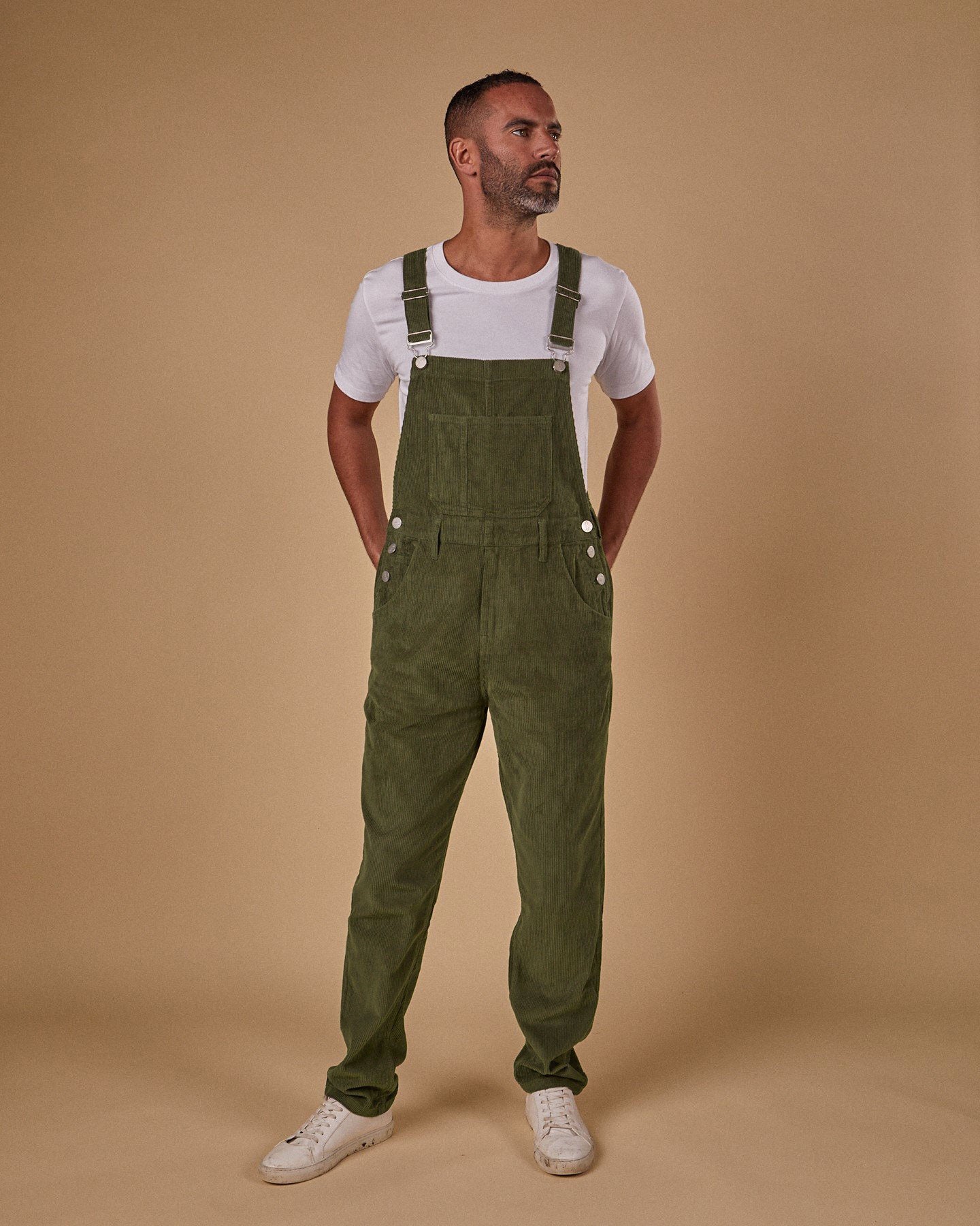Full-length, front view of model wearing olive-green corduroy dungarees with clear view of side button fastenings, front and bib pockets. 