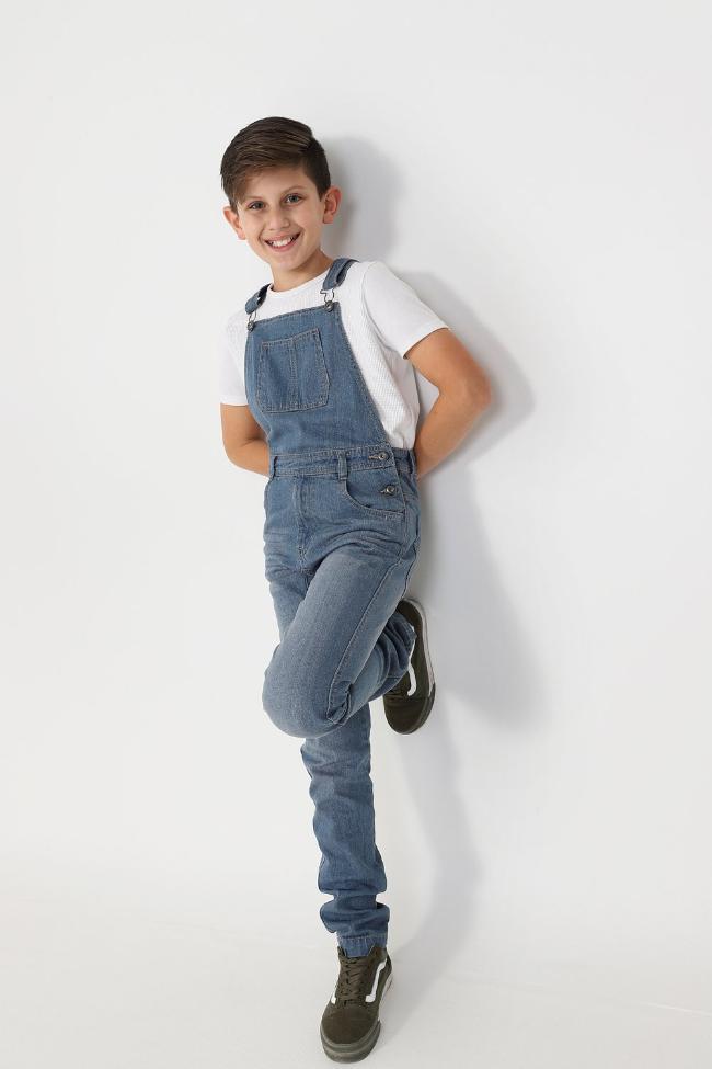 Full frontal leaning against wall with hands behind back, wearing boys light wash denim overalls.