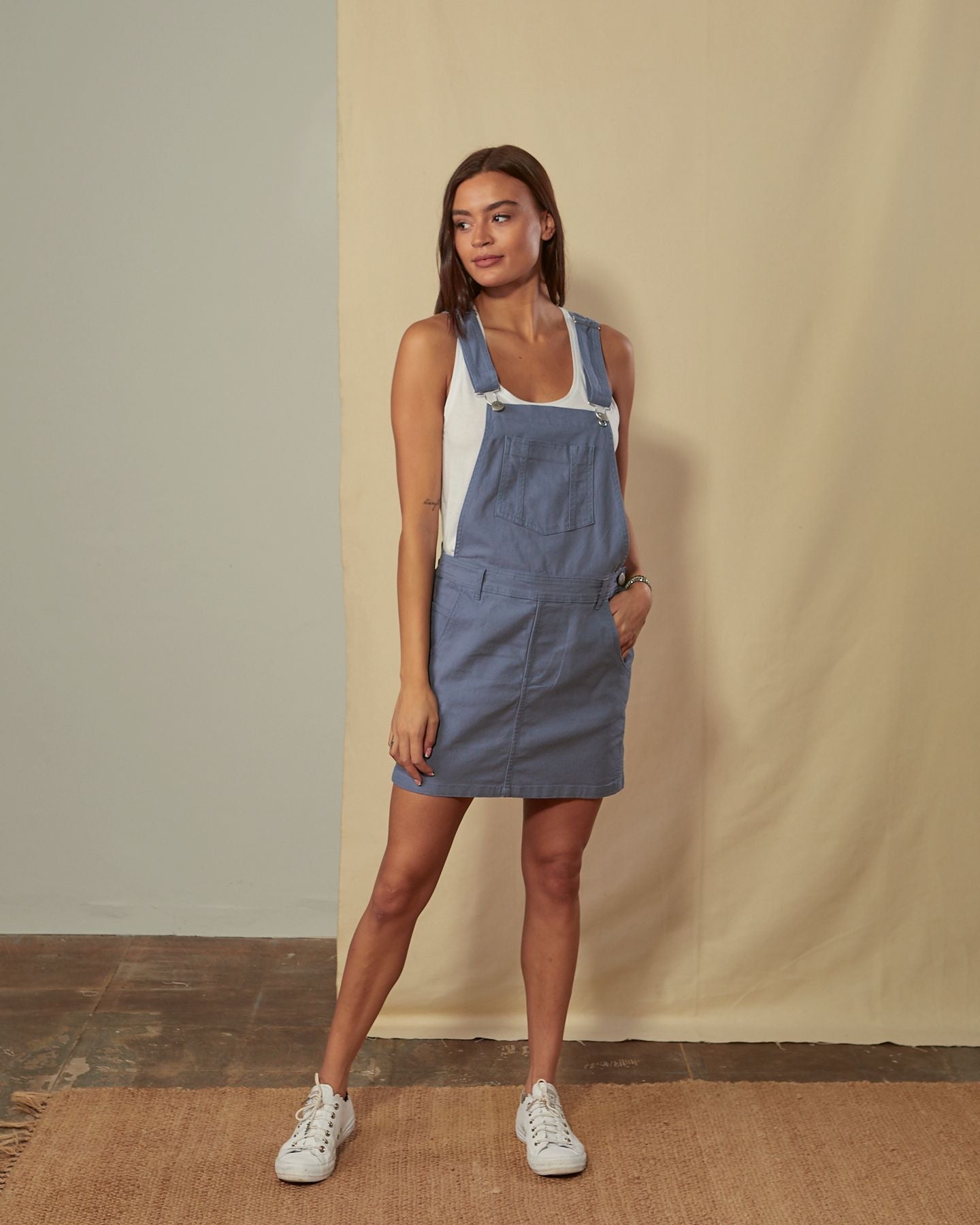 Full-length front view of model wearing sleeveless Cicely muted blue organic cotton dungaree-pinafore, showing belt loops and adjustable straps.
