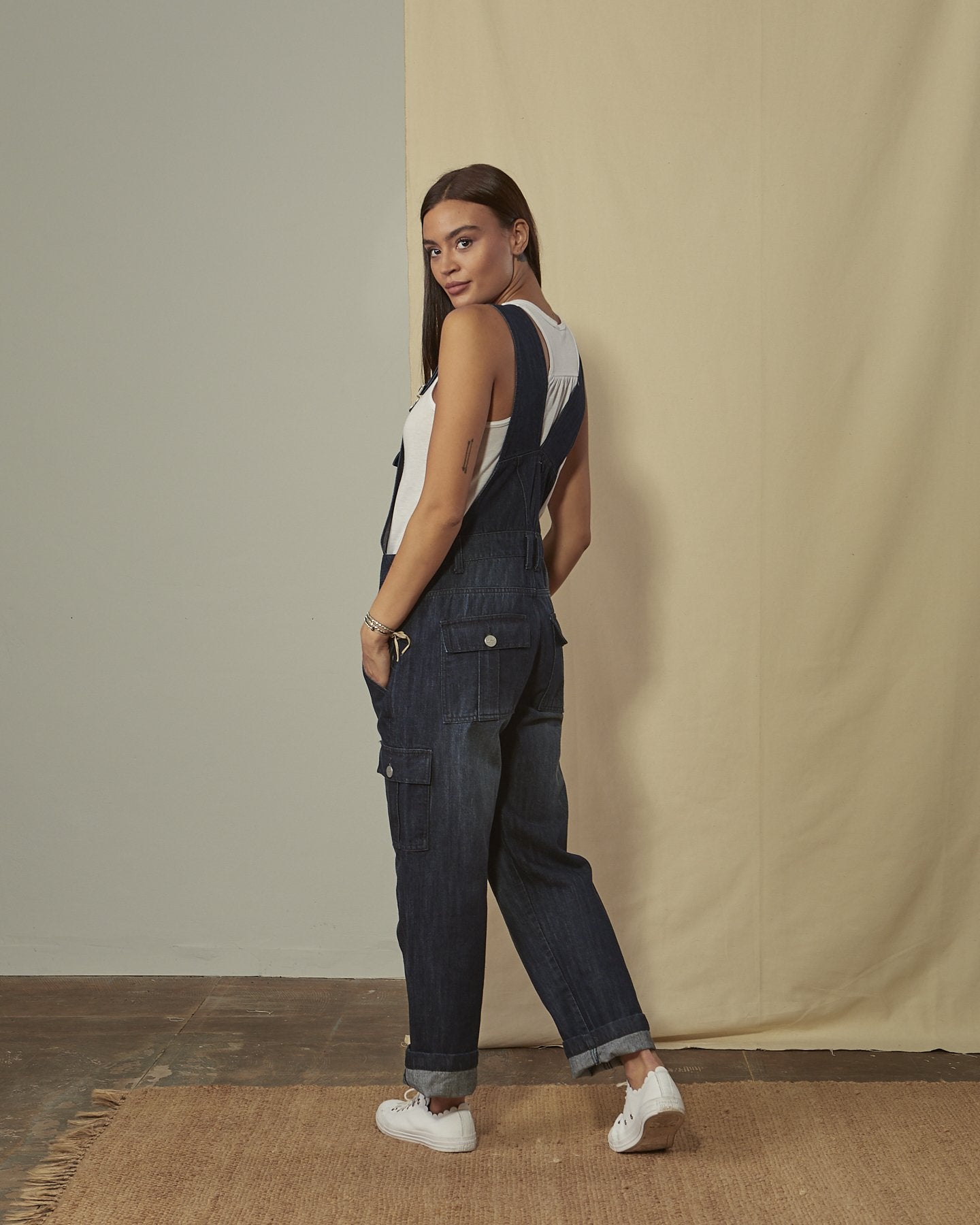 Back-side shot of women's darkwash denim cargo-pocket overalls with view of back button-down and cargo pockets.