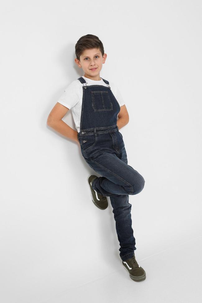 Full length pose leaning against wall wearing boys kids bib-overalls.