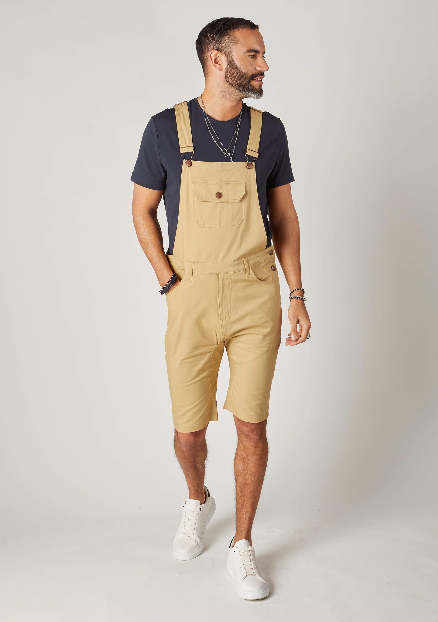 Full frontal walking pose wearing slim-fit, sand-coloured cotton bib-overall shorts from Dungarees Online. Hand in front-tight pocket with clear view of adjustable straps and large bib pocket.