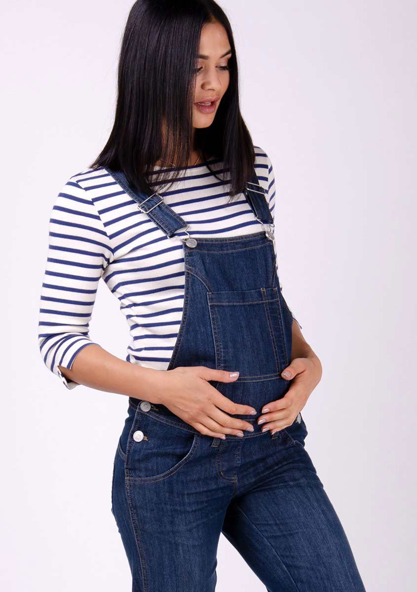 Two-thirds pose focussing on bib pocket, side buttons and adjustable straps of darkwash maternity overalls.