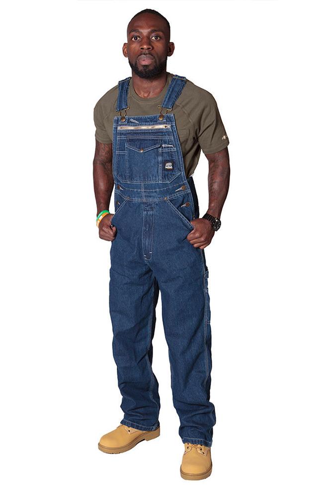 Full slightly angled frontal model pose showing men's denim bib overalls with utility pockets, adjustable straps and brass buckles.