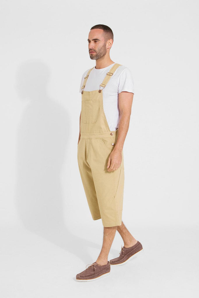 Full frontal pose angled to his right and walking, wearing bib-up sand coloured dungaree shorts from Dungarees Online.