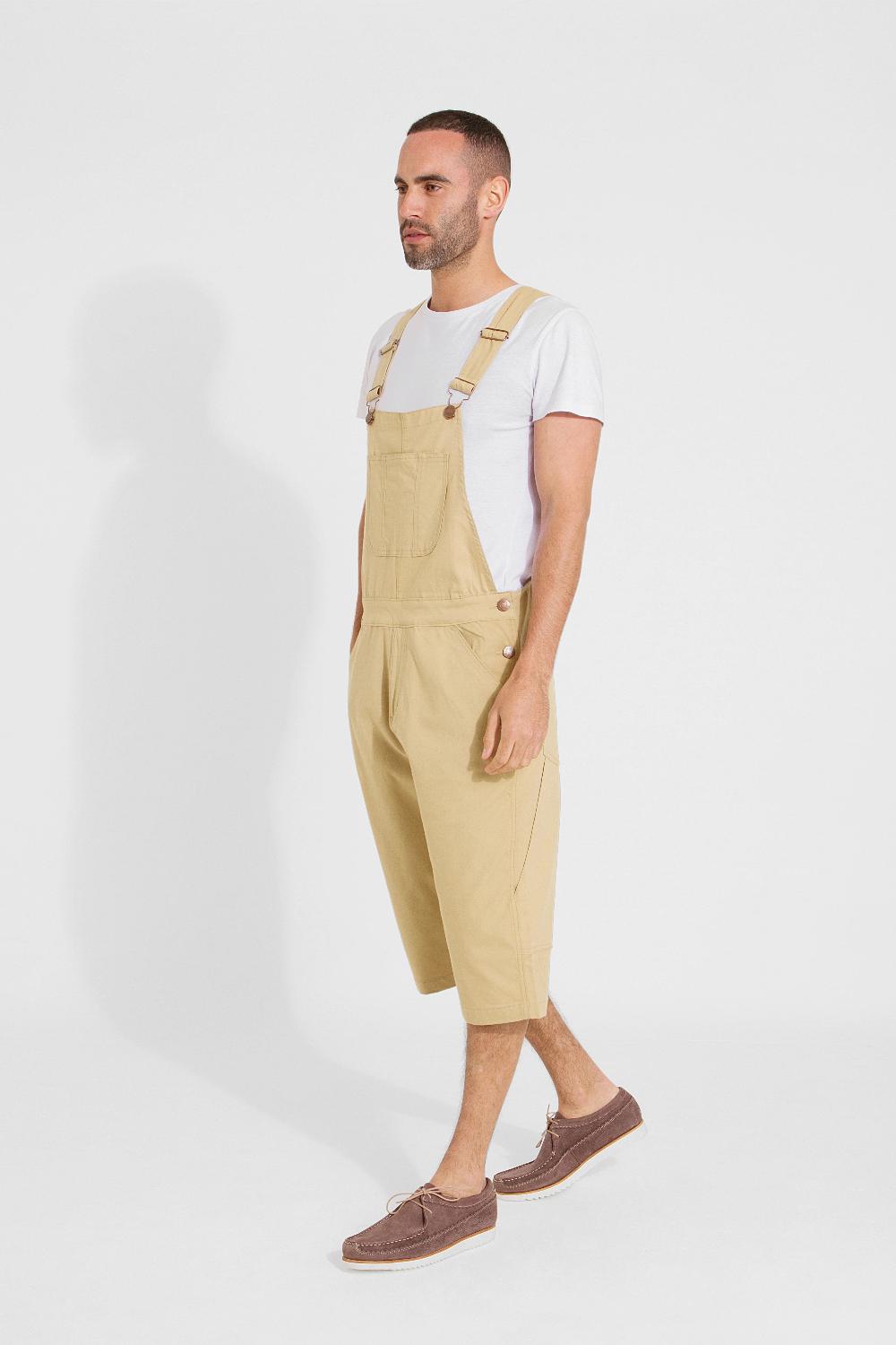 Full frontal pose angled to his right and walking, wearing bib-up sand coloured dungaree shorts from Dungarees Online.