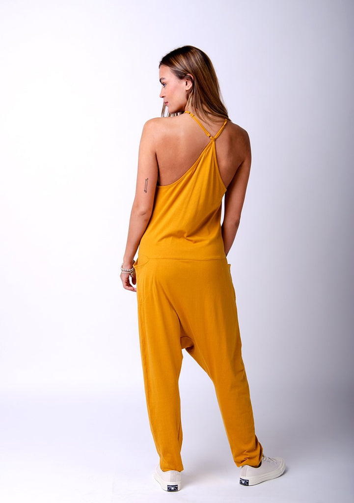 Full rear pose twisting to her left with hand in front pocket. Wearing Jools-style gold jersey jumpsuit showing crossed back straps.