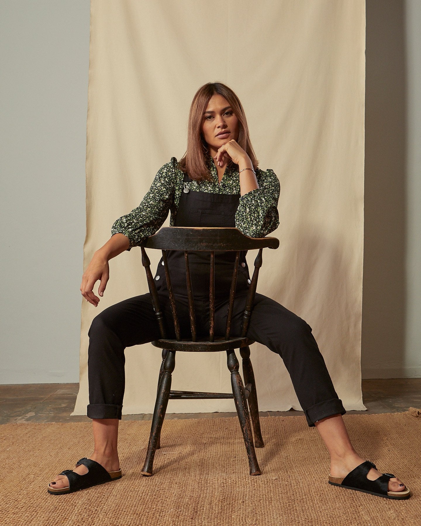 Sitting model wearing Harper black organic cotton dungarees with focus on button fastenings and turn-ups on tapered leg.