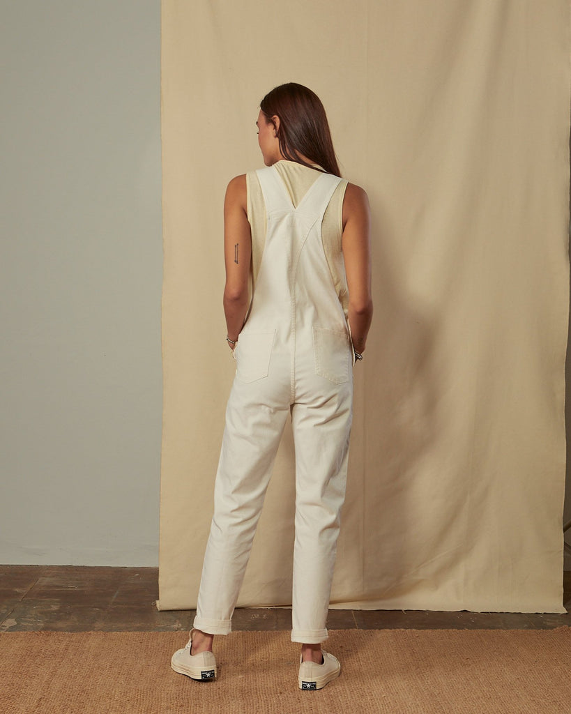Full-length side view of model wearing Harper cream organic cotton dungarees showing back pockets and tapered leg.