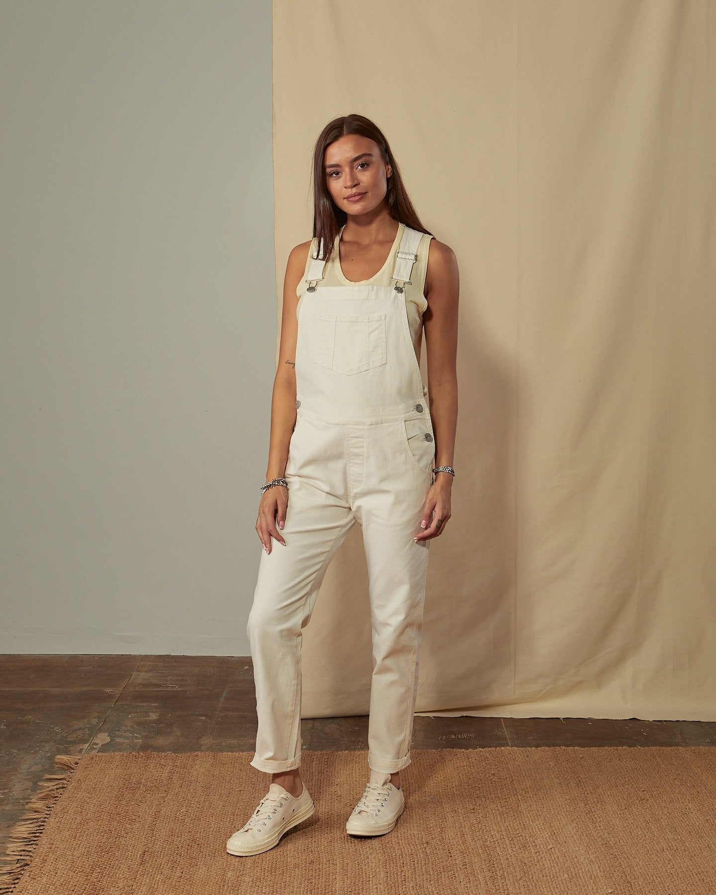 Full-length front view of model wearing Harper cream organic cotton dungarees with view of button fastening.