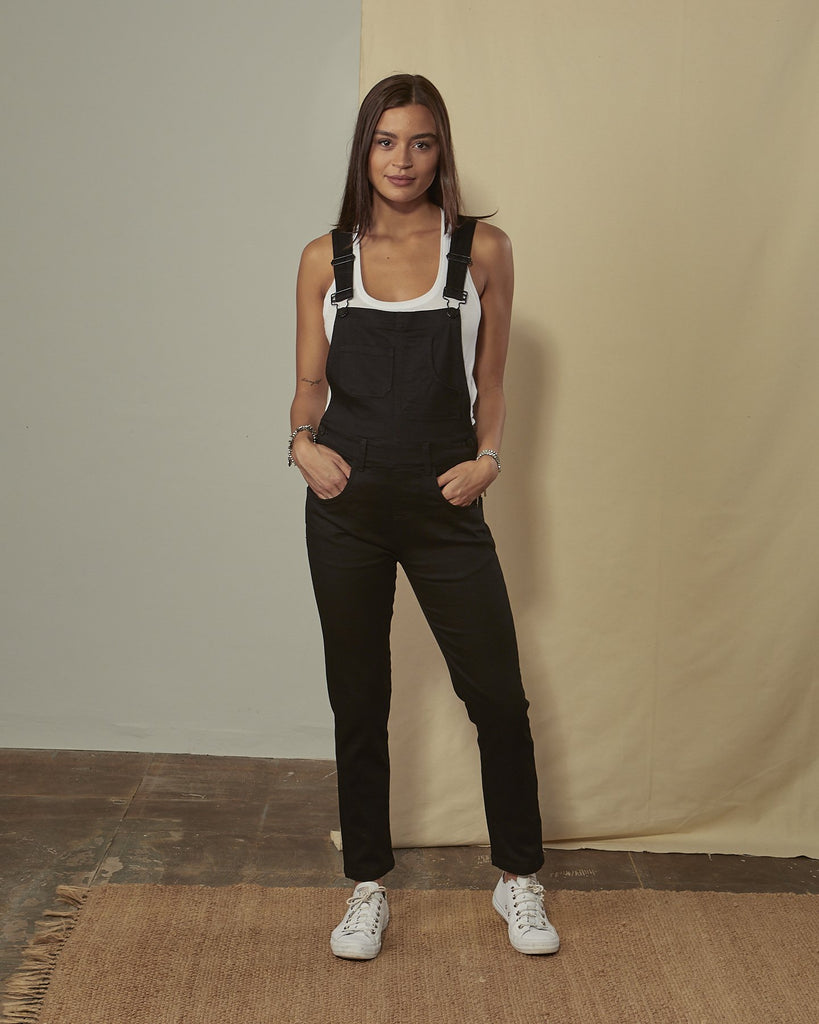 Full-length front view of model wearing Kate black cotton dungarees with hands in front pockets.