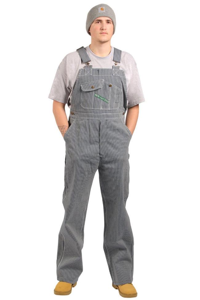 Full-length front view with hands in pockets, wearing ‘Key Apparel USA’ bib-overall made with premium materials and superior finish, paired with grey t-shirt and hat.