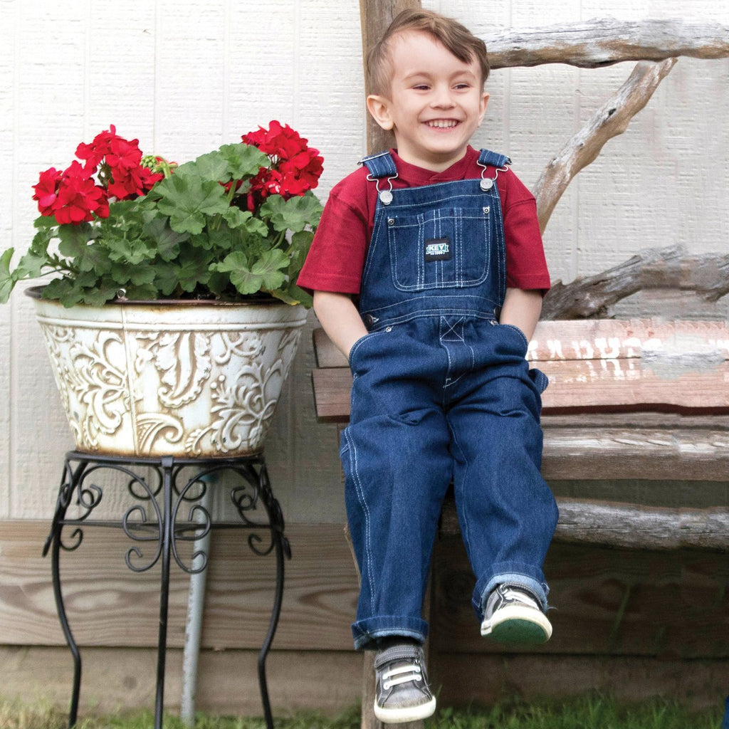 Young boy sat wearing dark blue denim dungarees from key industries USA with hands in front pockets.