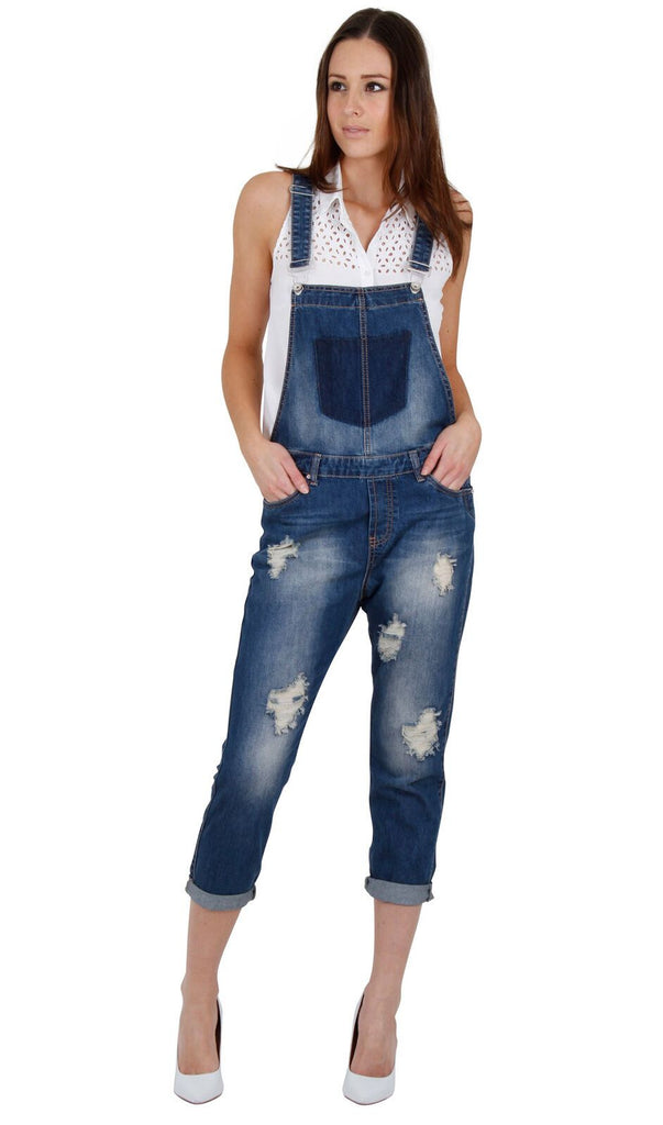 Full-length front view of model with hands in front pockets of women's destroyed denim dungarees.