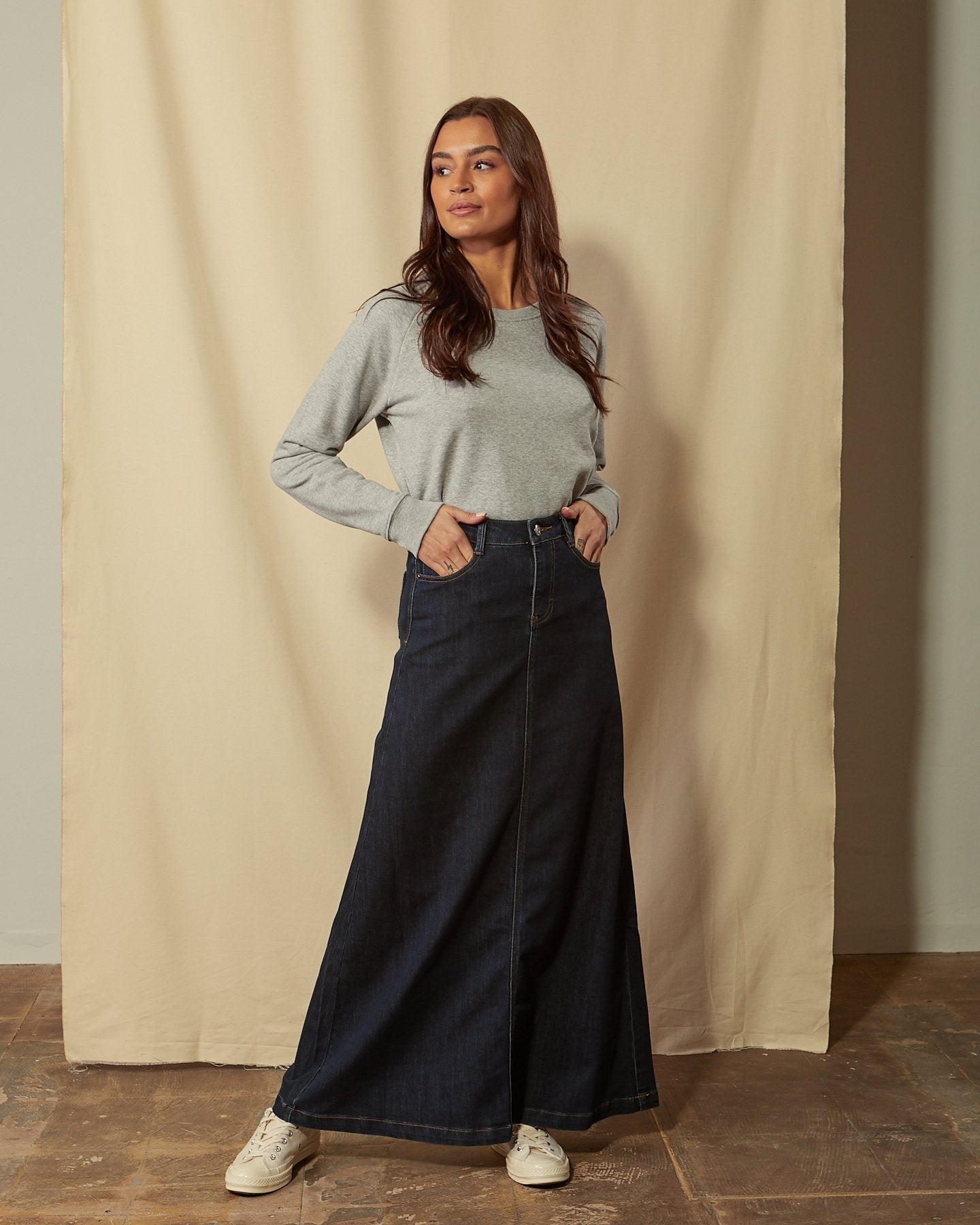 Full-length front view of model with hands in front pockets of dark blue A-line style denim skirt.