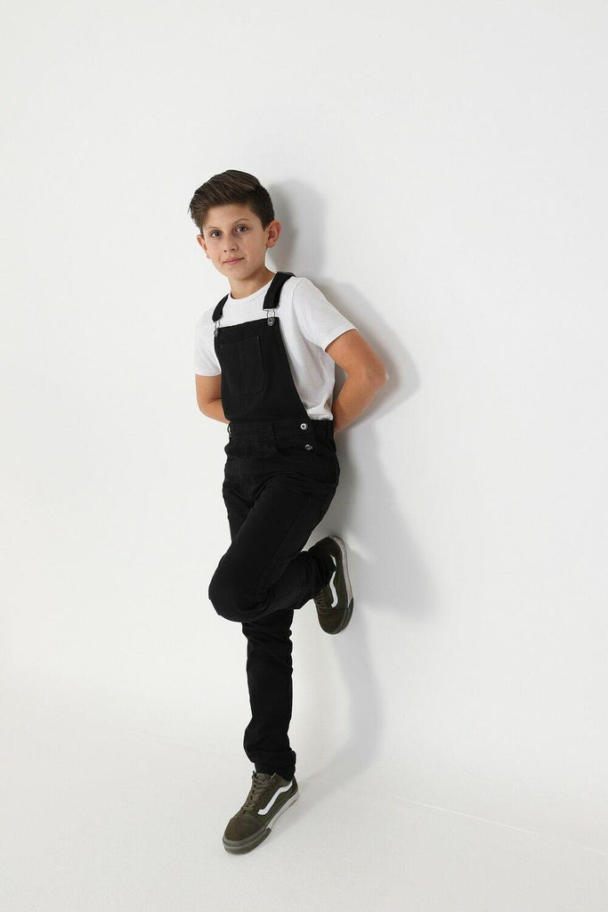 Full frontal leaning against wall with hands behind back, wearing boys black denim overalls.