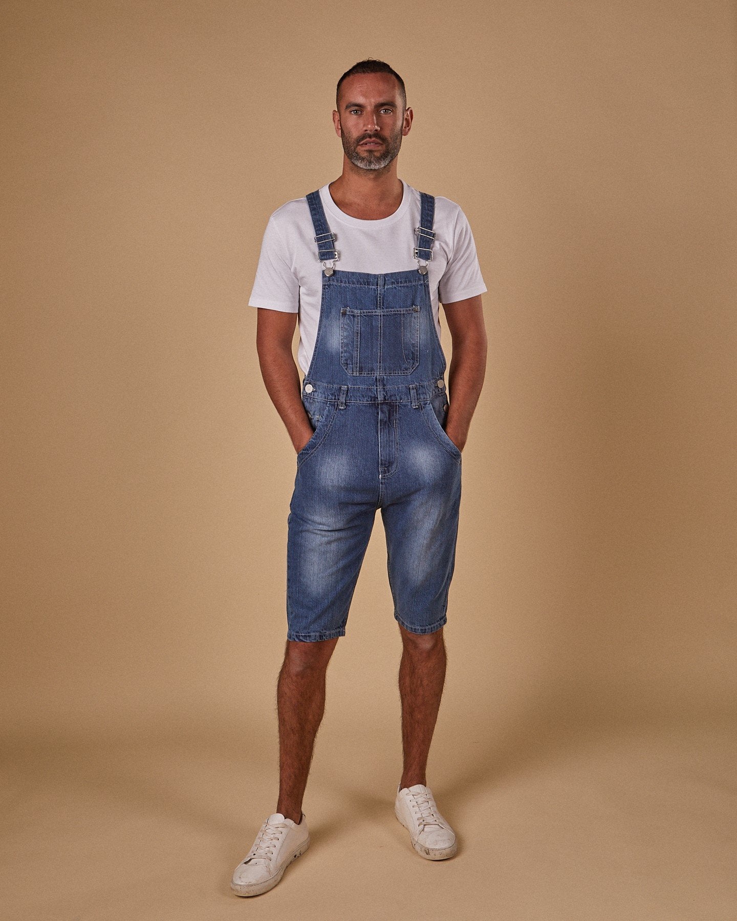 Full-length front view of model with hand in front pockets of Oliver denim dungaree shorts with view of adjustable straps and bib pocket.