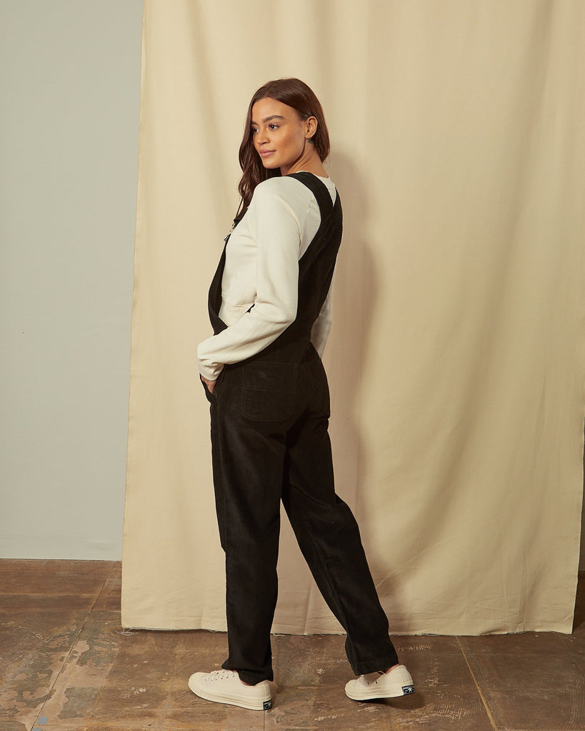 Full-length, slightly angled back view of model wearing Prue black corduroy dungarees with view of back pockets and straps.