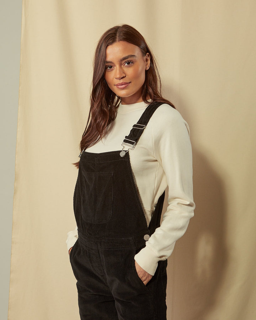 Slightly angled front view of the top-half of model with hands in front pockets of Prue black corduroy dungarees with view of adjustable buckle straps.