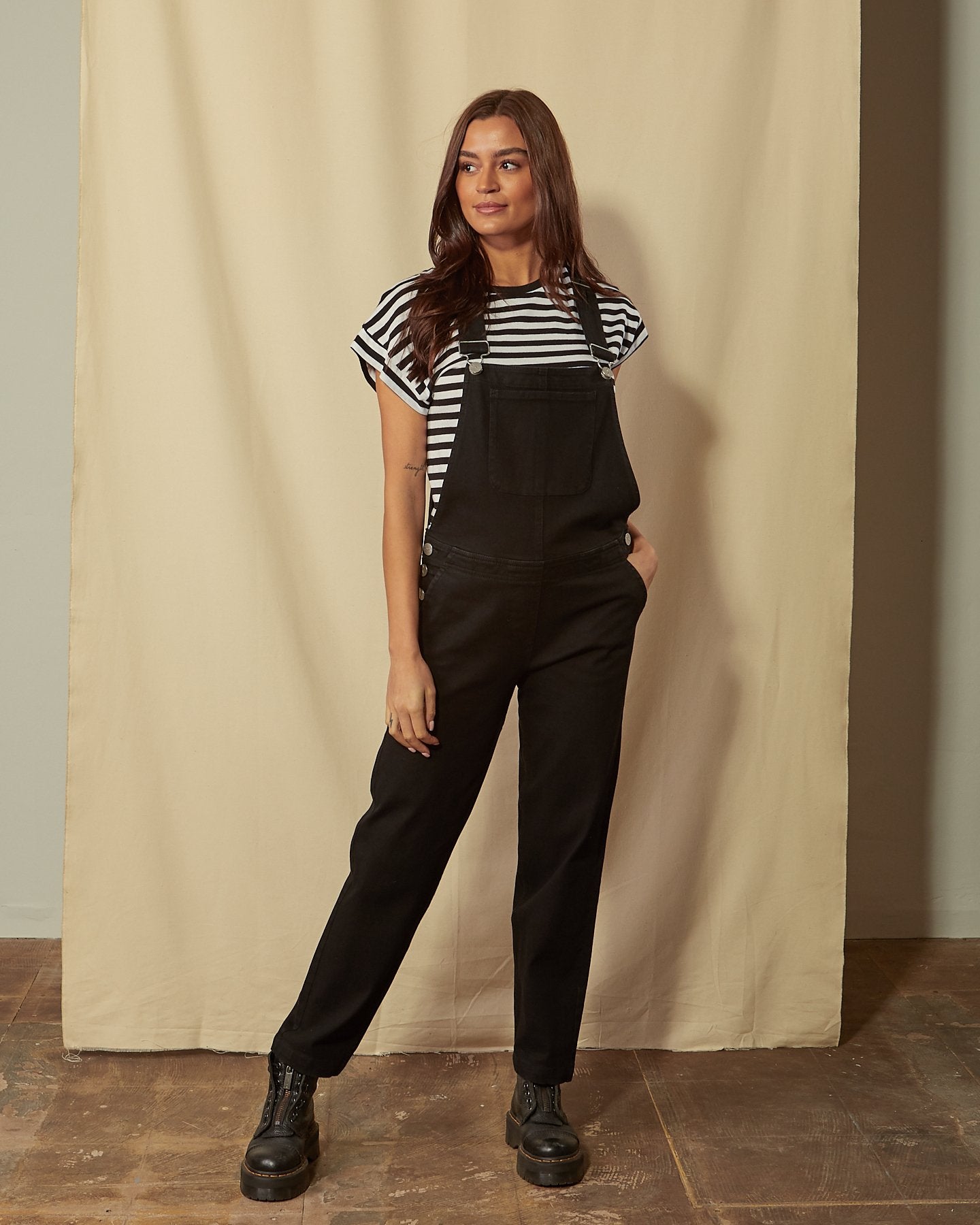 Full-length front view of model wearing Prue black cotton dungarees with hand in front pocket.