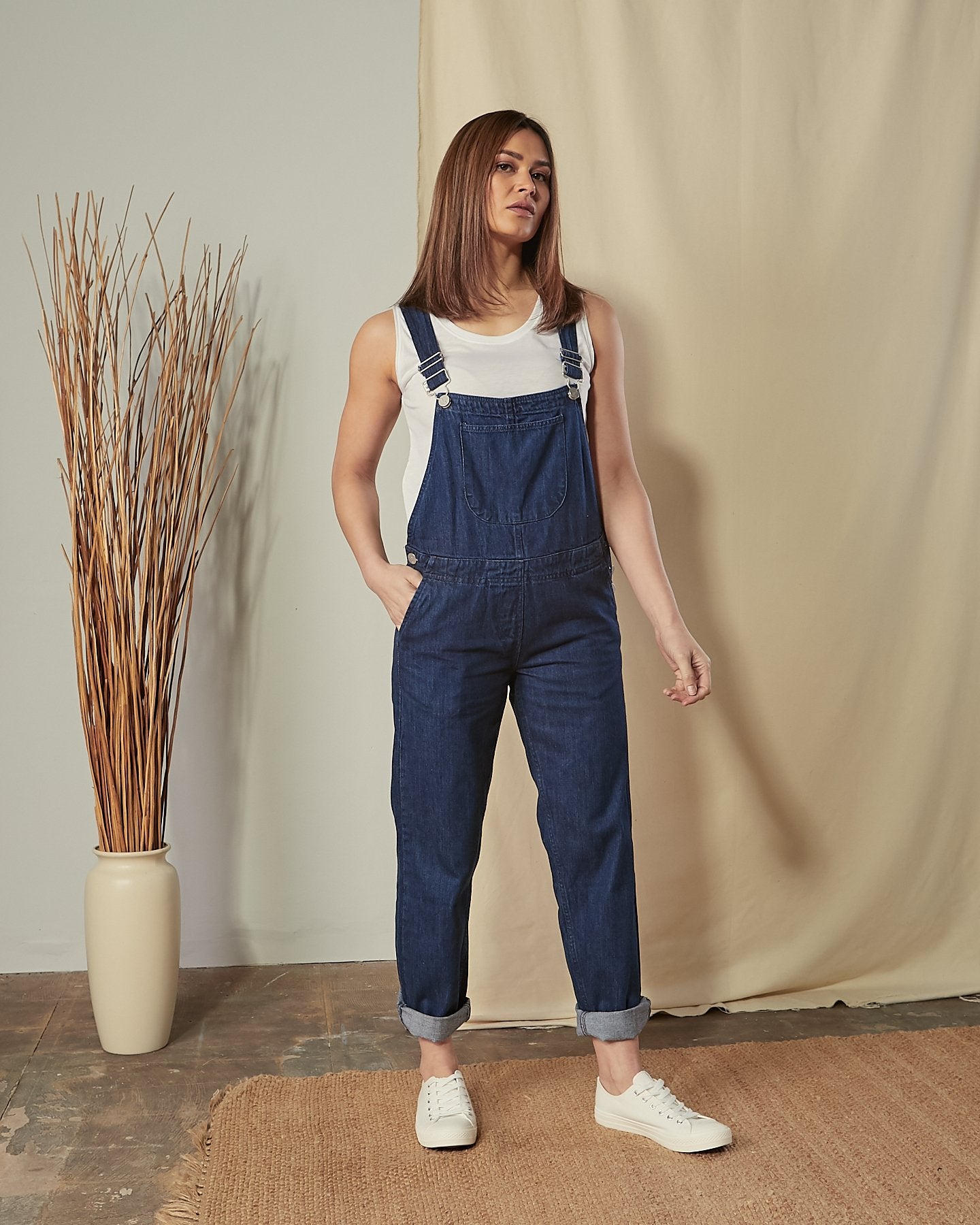 Full-length front view of model wearing Prue blue cotton dungarees with hand in front pocket.