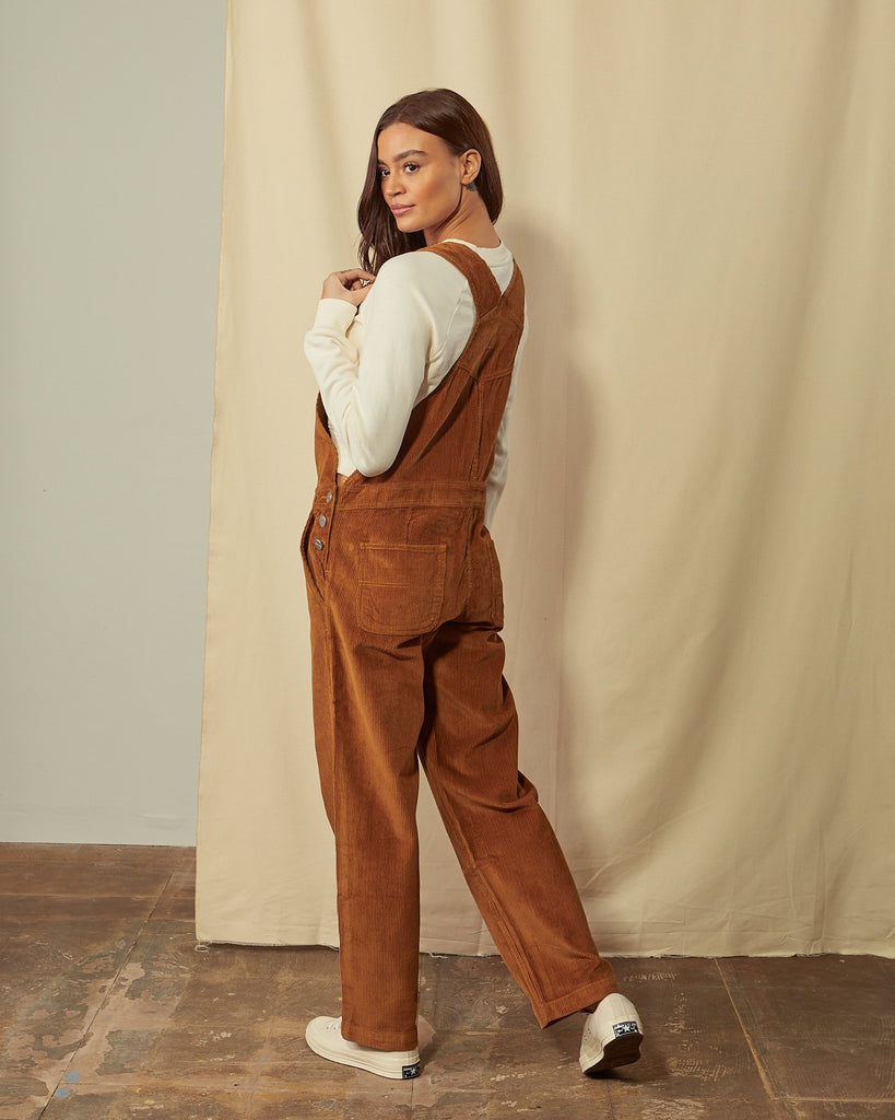 Full-length, slightly angled back view of model wearing Prue brown corduroy dungarees with view of 3-button side fastening, back pockets and straps.