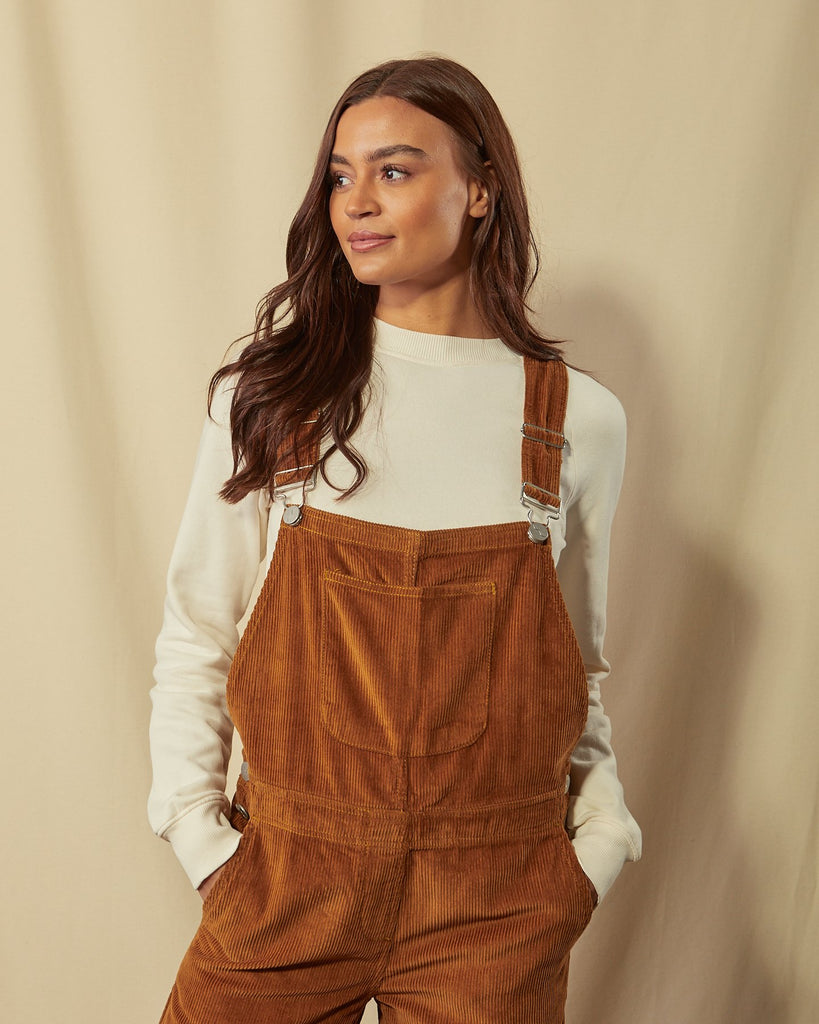 Slightly angled front view of the top-half of model with hands in front pockets of Prue brown corduroy dungarees with view of adjustable buckle straps.