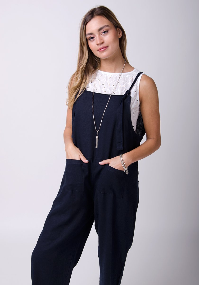 Two-thirds pose with hands in front pockets, wearing basic linen, dungaree style jumpsuit.