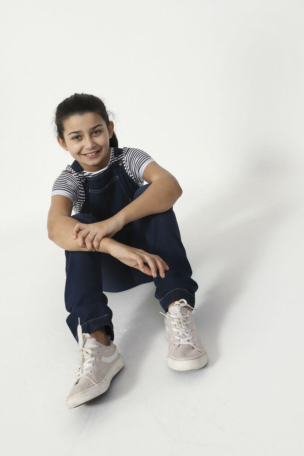 Girl facing ahead, sitting on floor with stretched legs, wearing slim fit denim dungarees with adjustable straps.